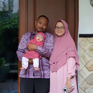 Une famille heureuse se tient debout, avec un père tenant un bébé et une mère souriante portant un hijab rose, posant devant une porte marron.