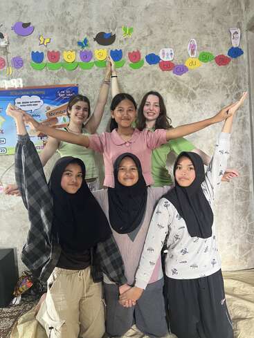 Six girls pose together indoors, smiling and creating a pyramid formation with their arms. Colorful educational decorations and posters are visible on the wall behind them.
