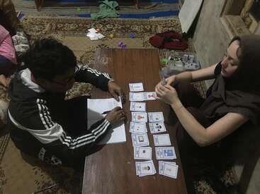 Two people sit on the floor around a low table, playing an educational card game, possibly language-based, with cards and a notebook, in a cozy room.