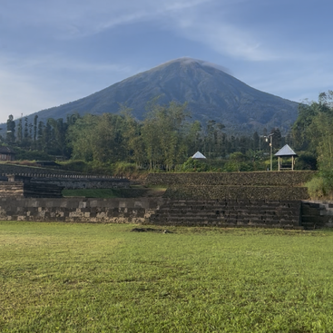 Ein majestätischer Vulkan erhebt sich unter einem klaren blauen Himmel, umgeben von üppigem Grün, alten Steinbauten, Treppen und friedlichen Pavillons inmitten einer ruhigen Landschaft.