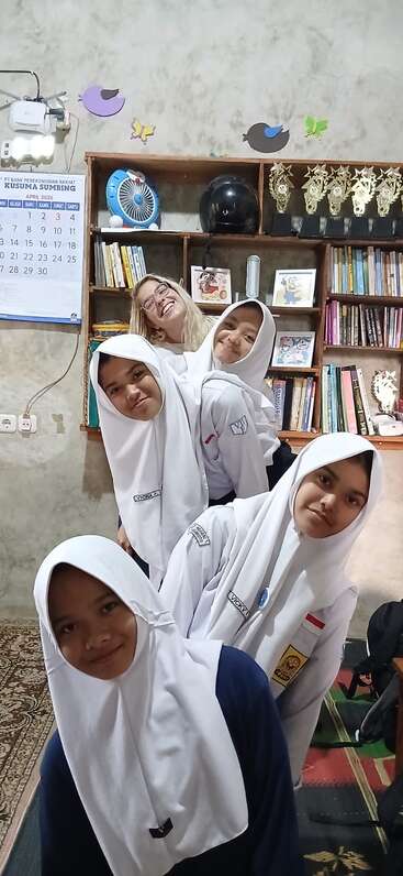 Cinco meninas sorridentes, quatro delas com hijabs brancos e uniformes escolares, posam juntas em um quarto aconchegante com estantes de livros, troféus, um calendário e decorações coloridas nas paredes.