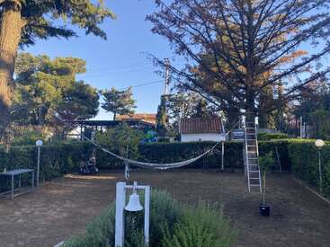 Uma cena tranquila de jardim com uma rede pendurada entre as árvores, um sino branco em primeiro plano, uma escada, vegetação, uma cerca viva e um pequeno galpão.