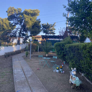 Uma cena de jardim com um caminho estreito de pedras, mesa e cadeiras verdes, pequenas estátuas decorativas, sebes bem aparadas, árvores e um céu azul claro ao fundo.