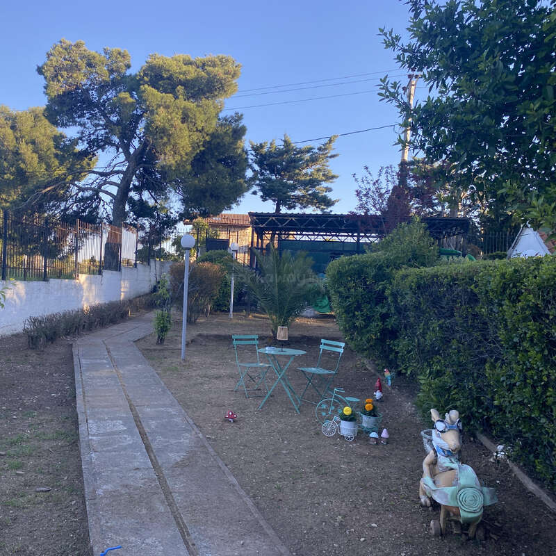 Uma cena de jardim com um caminho estreito de pedras, mesa e cadeiras verdes, pequenas estátuas decorativas, sebes bem aparadas, árvores e um céu azul claro ao fundo.