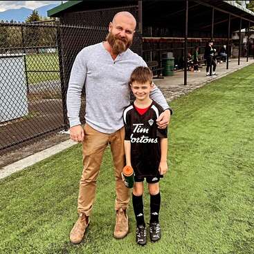 The image depicts a man and a boy standing on a soccer field, with the man\'s arm around the boy, both wearing casual attire, and the boy dressed in a soccer uniform.