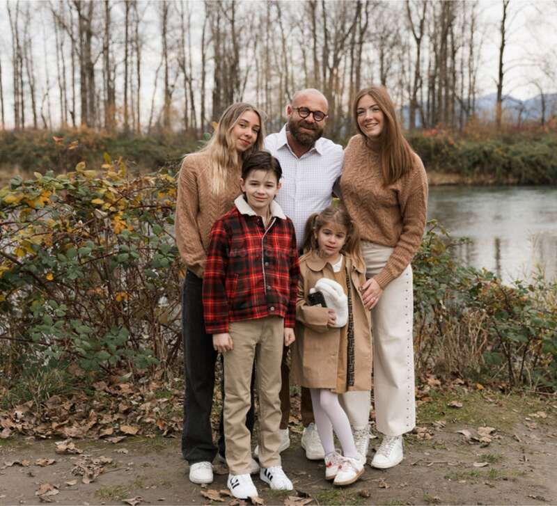 A family of five stands outdoors by a river. They wear autumn clothing and smile together, surrounded by bare trees and fallen leaves on the ground.