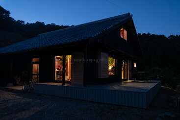 A cozy house with large windows glows warmly at dusk. Surrounded by trees, it sits peacefully with a bike nearby and an inviting outdoor deck.