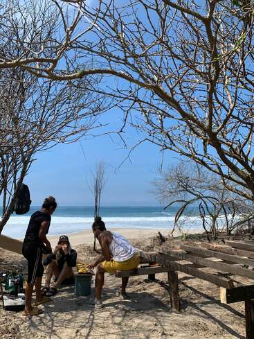 Three people relax and prepare food under leafless trees on a sandy beach, with wooden benches nearby and the ocean waves visible in the background under blue sky.