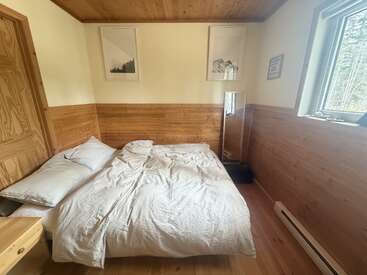 Este acogedor dormitorio presenta una decoración en madera, una cama sin hacer con sábanas blancas, cuadros con motivos naturales, un espejo, una ventana con vistas al bosque y luz natural.