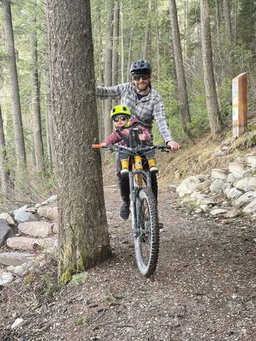 Un hombre y un niño, ambos con casco y gafas de sol, van en bicicleta de montaña por un sendero forestal. El hombre se sujeta a un árbol para mantener el equilibrio.