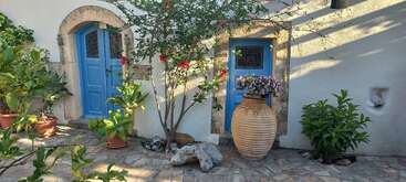 A charming courtyard features blue doors, potted plants, flowering bushes, and a large terracotta vase. Stones and lush greenery decorate the sunlit stone-paved area beautifully.