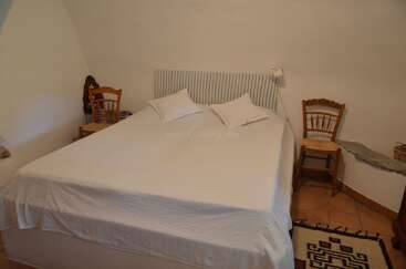 A cozy bedroom with a large bed covered in white linen and two pillows, wooden chairs, a radiator headboard, bedside tables, and tiled floor with rugs.