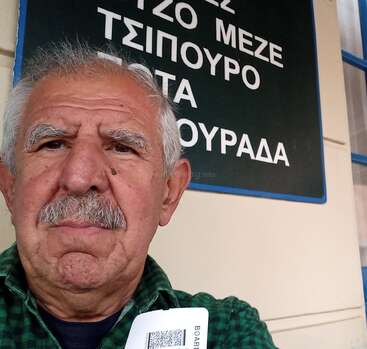 An older man with gray hair and mustache takes a selfie in front of a Greek sign, holding what appears to be a boarding pass or ticket.