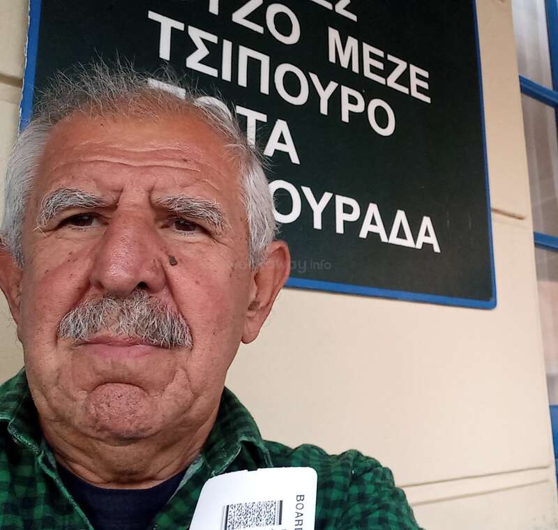 An older man with gray hair and mustache takes a selfie in front of a Greek sign, holding what appears to be a boarding pass or ticket.