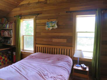 This cozy wooden bedroom features a bed with a pink cover, two large windows, green curtains, a lamp, bookshelves, and soft natural daylight.
