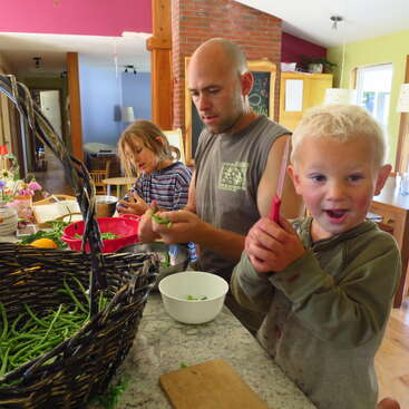 A man and two children are preparing green beans together in a cozy kitchen. The youngest child looks excited, holding a knife, smiling playfully.