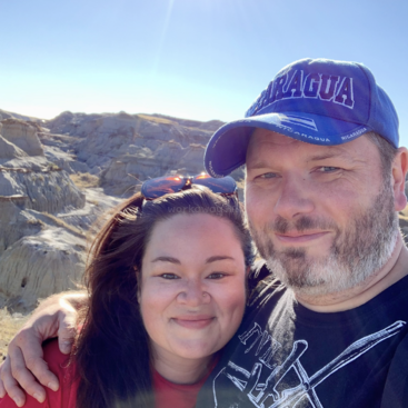 L'image montre un homme et une femme posant pour un selfie dans un paysage désertique avec des collines rocheuses et un ciel bleu clair, tous deux portant des vêtements décontractés et souriant.