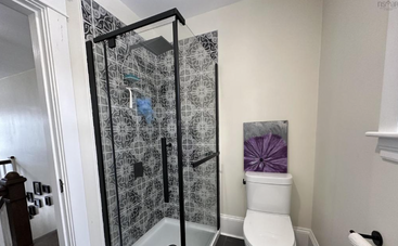 Cette salle de bain comprend une douche vitrée avec des carreaux à motifs, des toilettes blanches, des murs clairs, une œuvre d'art représentant des fleurs violettes et une atmosphère moderne et lumineuse.