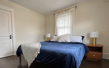 Cette chambre confortable est équipée d'un couvre-lit bleu, de trois oreillers, de deux lampes dorées posées sur des tables de chevet en bois, d'une porte blanche, d'un parquet et de rideaux transparents à la fenêtre.