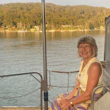 Eine lächelnde Frau sitzt in einem Boot an einem ruhigen Fluss, im Hintergrund bietet sich ein malerischer Ausblick auf Bäume, Häuser am Hang und Yachten auf der anderen Seite des Wassers.