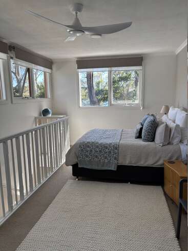 Ein helles, gemütliches Schlafzimmer mit einem ordentlich gemachten Bett mit gemusterten Akzenten, großen Fenstern, die viel Tageslicht hereinlassen, einem Deckenventilator und einer minimalistischen Einrichtung in neutralen Farbtönen.