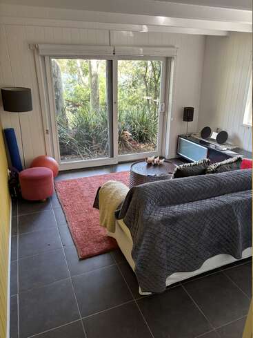 Ein gemütliches Wohnzimmer mit einem weißen Sofa, grauen und beigen Überwürfen, einem roten Teppich, einem Gymnastikball, einem Fernseher, einer Stehlampe, Schiebetüren und einem Blick ins üppig grüne Grün draußen.