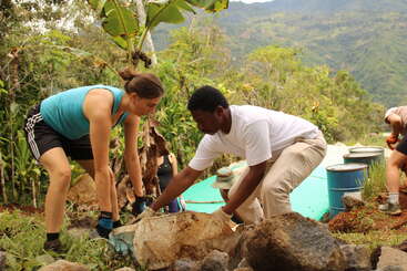 Duas pessoas estão trabalhando juntas ao ar livre, levantando pedras em um ambiente exuberante e montanhoso. Elas parecem concentradas, colaborando em um projeto com outras pessoas próximas, cercadas por vegetação.