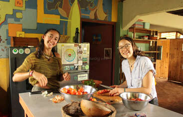 Duas mulheres estão sorrindo e preparando comida juntas em uma cozinha colorida. Há legumes frescos e tigelas sobre o balcão. A atmosfera é alegre e convidativa.