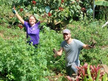Two people joyfully gardening, surrounded by lush green plants and colorful flowers. Both are smiling, wearing gloves, and appear to be harvesting or weeding.