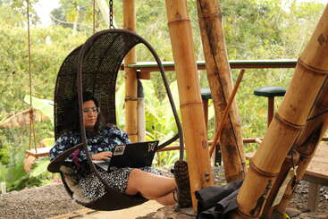 A woman sits in a hanging chair, working on her laptop, surrounded by bamboo and lush greenery, creating a relaxed outdoor workspace with a tropical vibe.