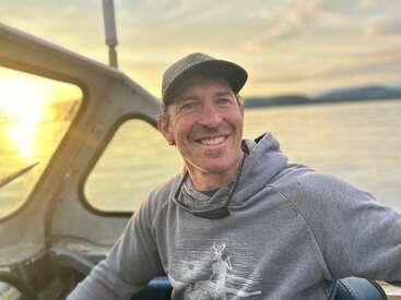 Ein lächelnder Mann in Kapuzenpulli und Mütze sitzt bei Sonnenuntergang in einem Boot, mit ruhigem Wasser und Bergen im Hintergrund, und genießt einen friedlichen Moment.
