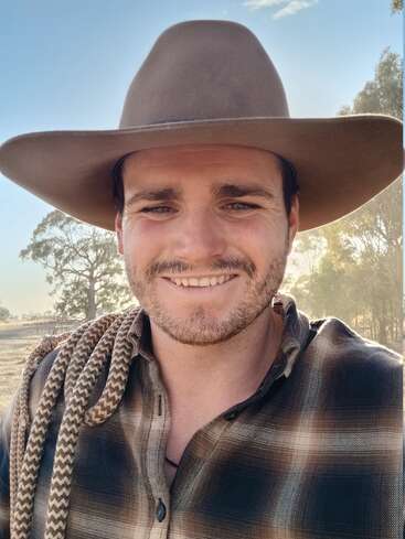 A young man wearing a wide-brimmed hat and plaid shirt smiles outdoors, holding a rope over his shoulder. Trees and blue sky are visible behind him.