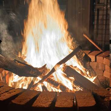 A roaring bonfire burns brightly within a circular brick fire pit, logs stacked in the flames. Warm, golden firelight illuminates the surrounding area at night.