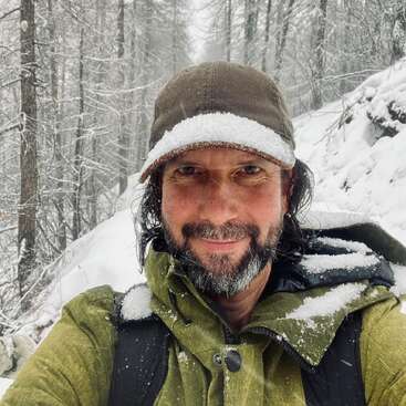 A man with a beard and mustache, wearing a green jacket and cap, stands smiling in a snowy forest, surrounded by trees covered in fresh snow.