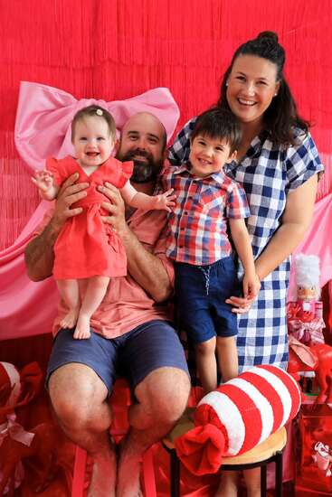 Uma família feliz de quatro pessoas posa, sorrindo em um cenário vermelho e rosa vibrante com decorações festivas, incluindo um laço gigante e um adereço de doces, criando uma atmosfera alegre.