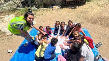 In this image, a group of children sit together on a blue tarp outdoors, smiling, with their hands reaching towards the center. An adult supervises enthusiastically.