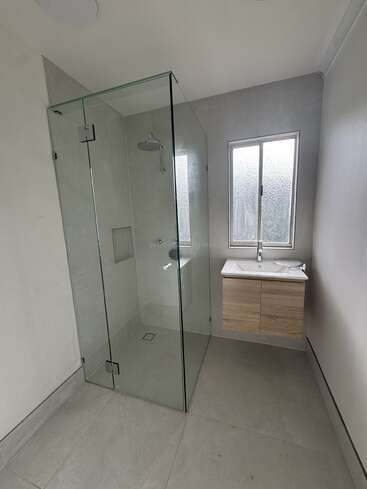 This modern bathroom features a frameless glass shower, rainfall showerhead, large gray tiles, a floating wooden vanity with sink, and a frosted window for privacy.