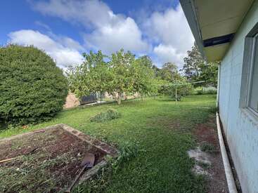 A backyard with green grass, a garden bed, a shovel, several trees, a rotary clothesline, bushes, a house wall, and a partly cloudy blue sky.