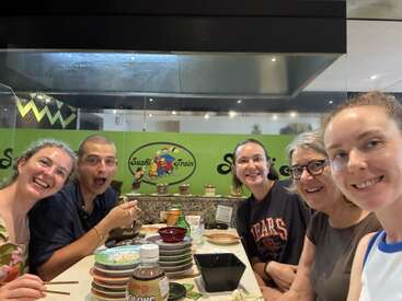 Cinco pessoas sorridentes sentam-se juntas em uma mesa de restaurante de sushi com pratos empilhados, pauzinhos e bebidas, desfrutando de uma refeição divertida no "Sushi Train".