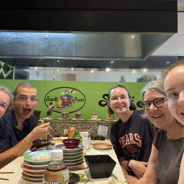 Five smiling people sit together at a sushi restaurant table with stacked plates, chopsticks, and drinks, enjoying a fun meal at "Sushi Train."