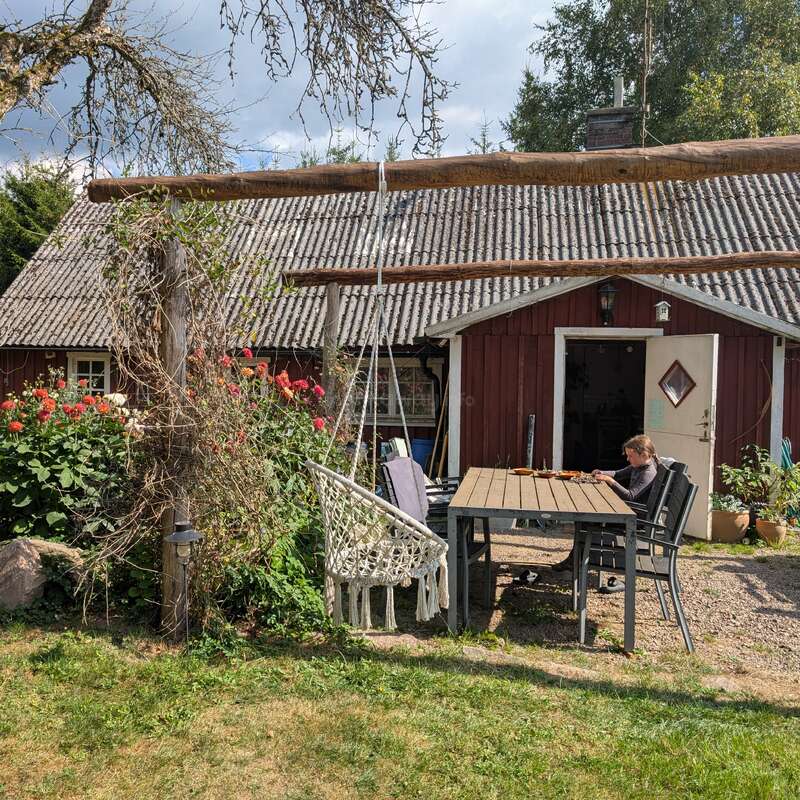 Uma casa de campo vermelha rústica com telhado de papelão ondulado, flores de jardim, um balanço de macramê, uma mesa ao ar livre e uma pessoa sentada sozinha, lendo ou trabalhando pacificamente do lado de fora.