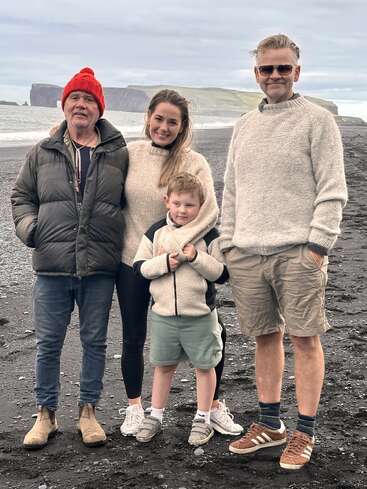 Eine Gruppe von vier Personen, darunter ein Kind, steht gemeinsam an einem schwarzen Sandstrand. Sie tragen warme Kleidung, lächeln und haben hinter sich Klippen und das Meer.