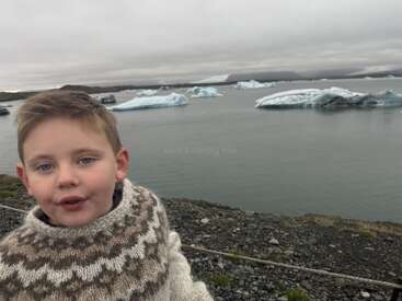 Ein kleines Kind in einem gemusterten Wollpullover steht an einem felsigen Ufer, während große Eisberge in einer ruhigen, grauen und bewölkten arktischen Landschaft treiben.