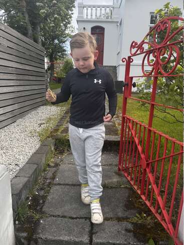 Ein kleiner Junge in einer schwarzen Jacke und grauen Jogginghosen geht mit Stöcken in der Hand an einem roten Tor vorbei, vor einem weißen Haus, das von üppigem Grün umgeben ist.