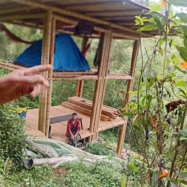 Uma estrutura de bambu fica em uma floresta verde e exuberante. Uma pessoa senta-se no nível inferior, enquanto uma tenda azul é montada acima. Gestos com as mãos.