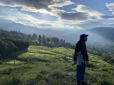 A person stands on a grassy hill, holding a blanket, overlooking a lush green valley with dramatic clouds and sunlight streaming through, surrounded by distant mountains.