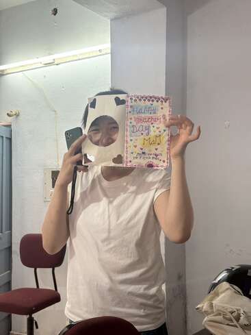 Una persona con camisa blanca sostiene una tarjeta decorada del Día de los Docentes y un teléfono, y se asoma juguetonamente a través de un recorte de papel con formas de corazón.