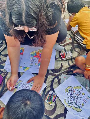 Una mujer ayuda a unos niños a colorear mariposas con lápices de colores. Están sentados en una alfombra estampada, concentrados en su actividad creativa. El ambiente parece relajado y educativo.