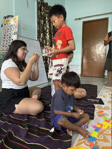 Tres niños y una mujer están sentados en el suelo, realizando actividades educativas y lúdicas. Hay tablas de aprendizaje, una pizarra blanca y piezas de puzzle de colores.