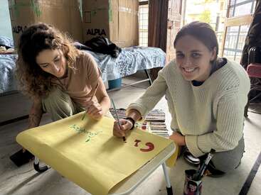 Dos mujeres se sientan en el suelo, sonrientes y trabajando juntas en un cartel de colores. La habitación es acogedora, con camas y cajas al fondo. Ambiente creativo.